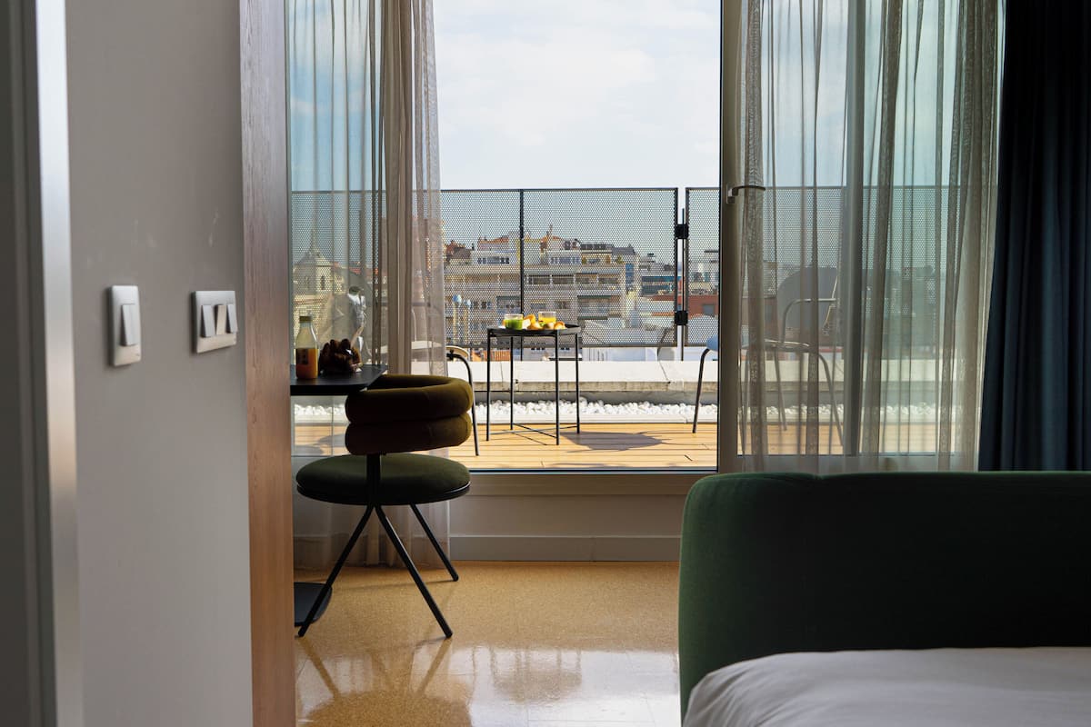 A hotel room with a large window, a green chair, a small table with fruits, and a bed.