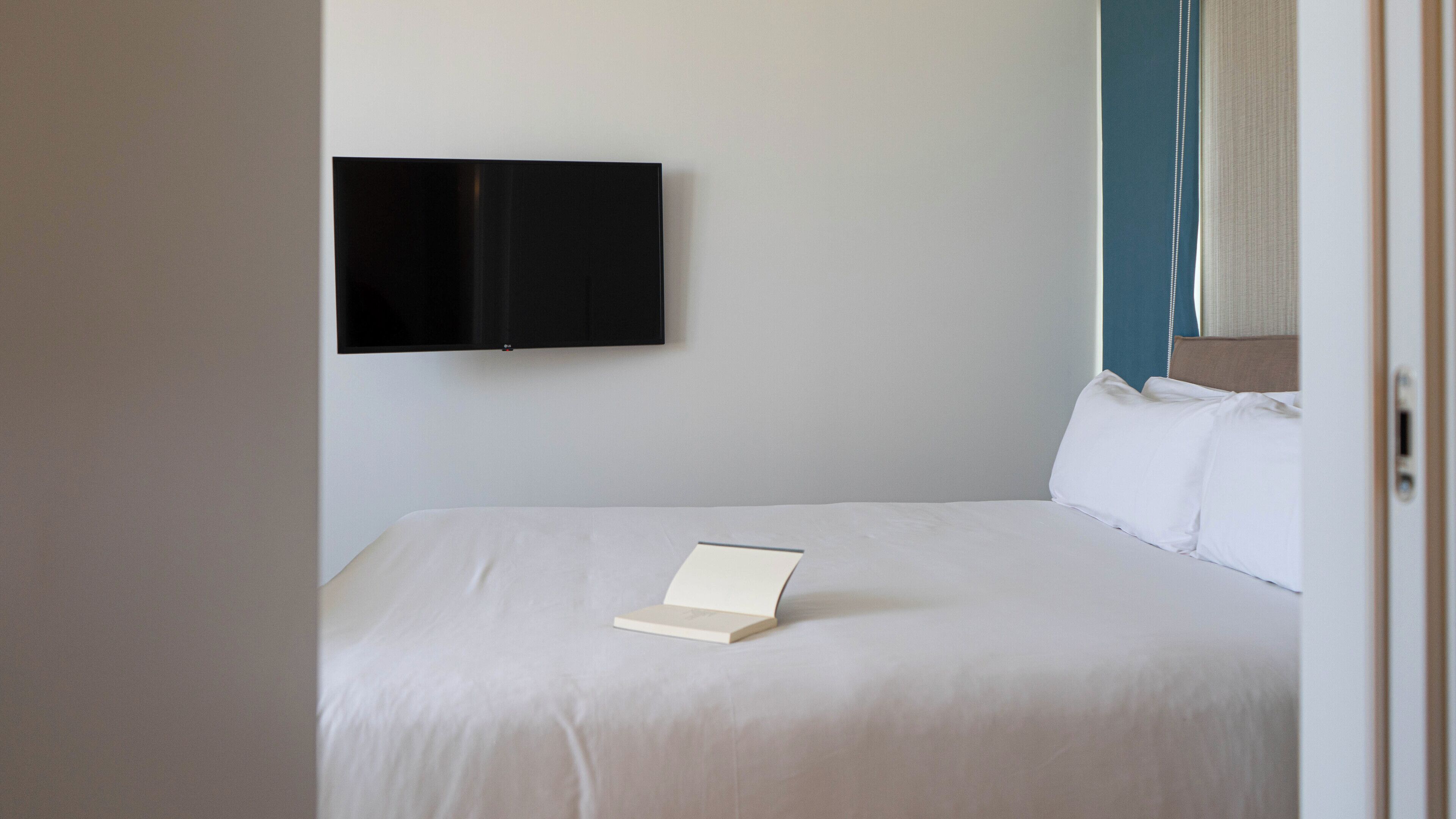 Una habitación de hotel con una cama, un televisor de pantalla plana montado en la pared y un libro sobre la cama.