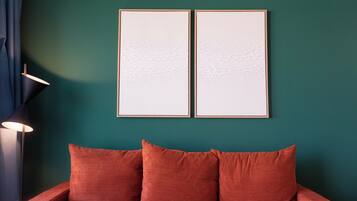 A room with a teal wall, two framed artworks, a red sofa with cushions, and a floor lamp.