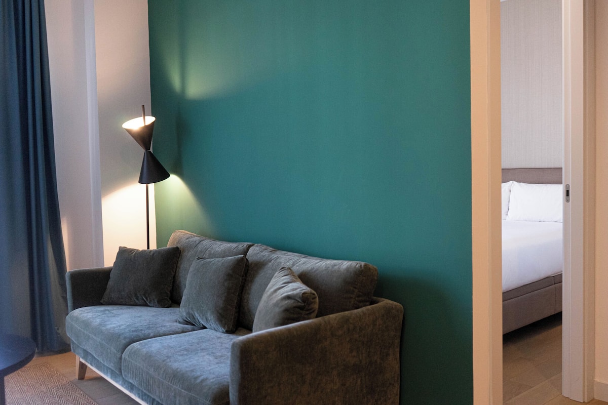 A room with a grey sofa, a floor lamp, and a bed with white bedding.