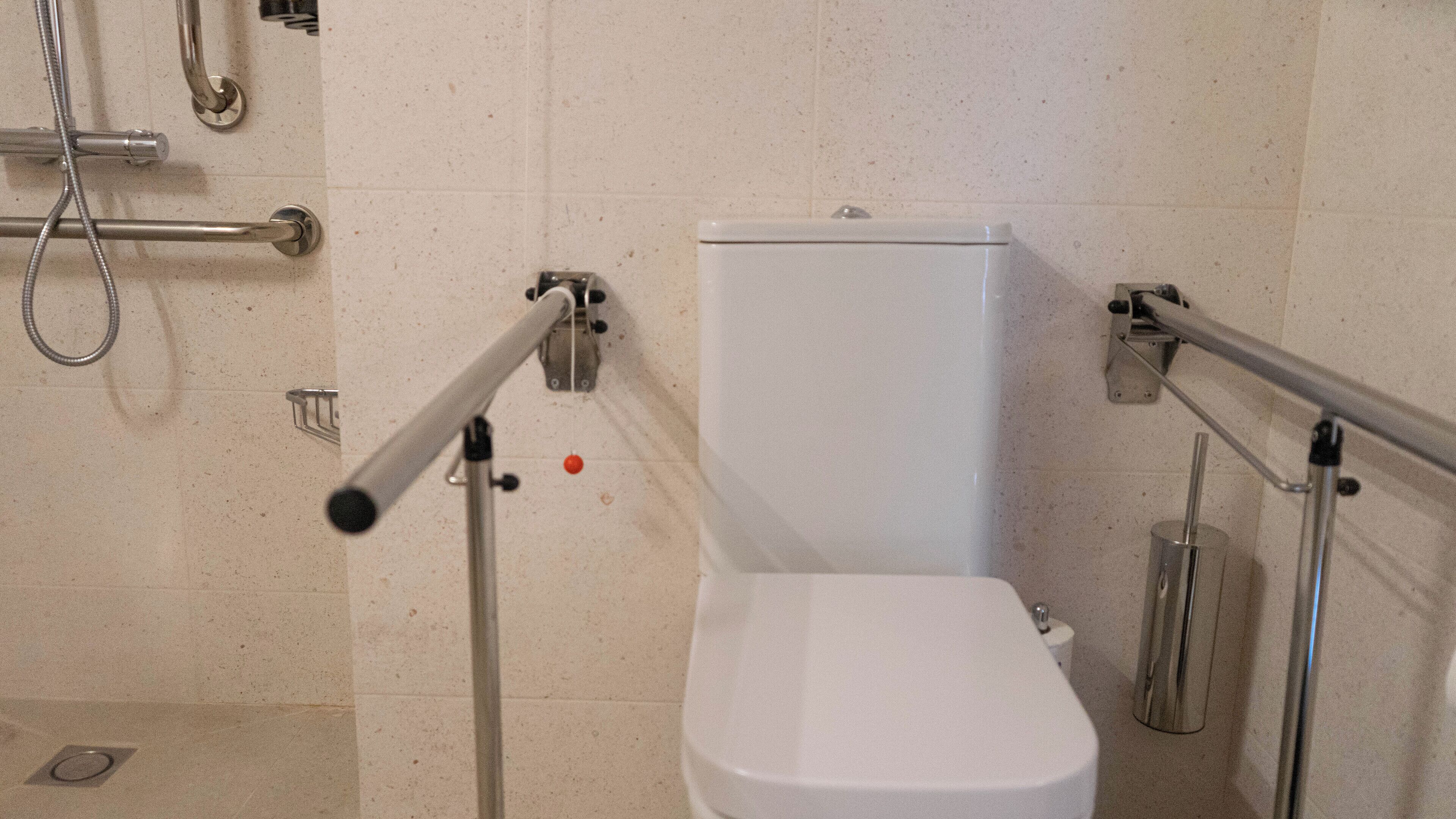 Kylpyhuone, jossa on valkoinen wc-istuin, tukikaiteet ja suihkutila.