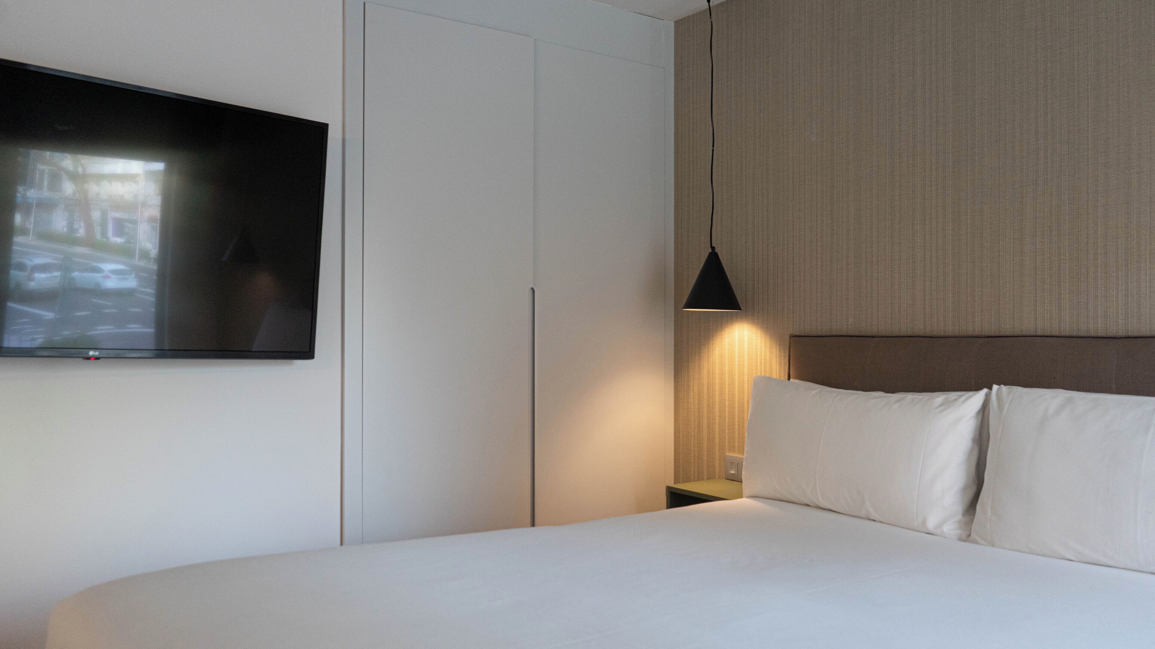 Habitación de hotel con una cama grande, un televisor de pantalla plana montado en la pared, una mesita de noche con lámpara y cabecero.