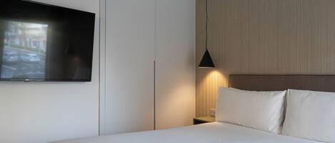 Habitación de hotel con una cama grande, un televisor de pantalla plana montado en la pared, una mesita de noche con lámpara y cabecero.