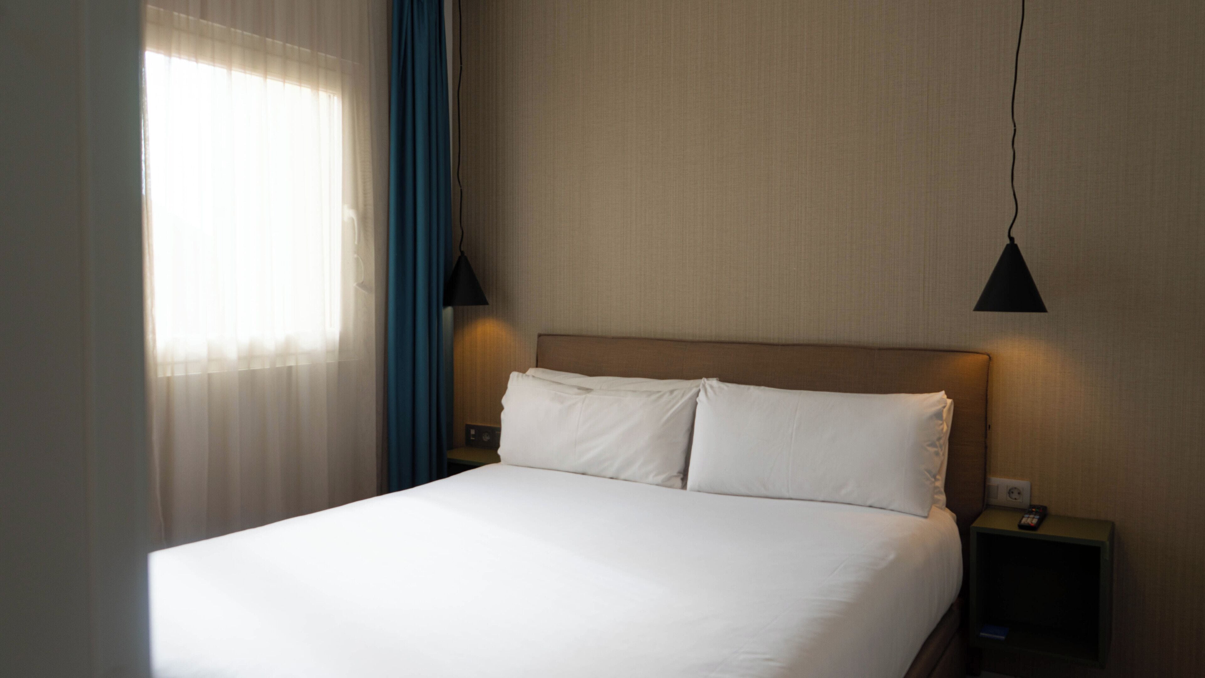 A neatly made bed in a hotel room with a headboard, two bedside tables, a lamp, and a window with curtains.