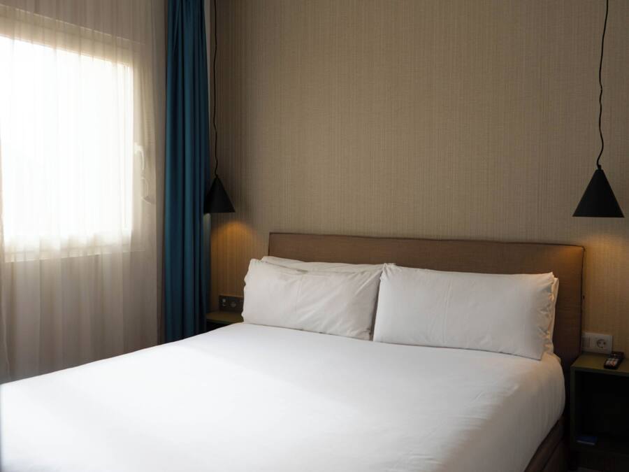 A neatly made bed in a hotel room with a headboard, two bedside tables, a lamp, and a window with curtains.