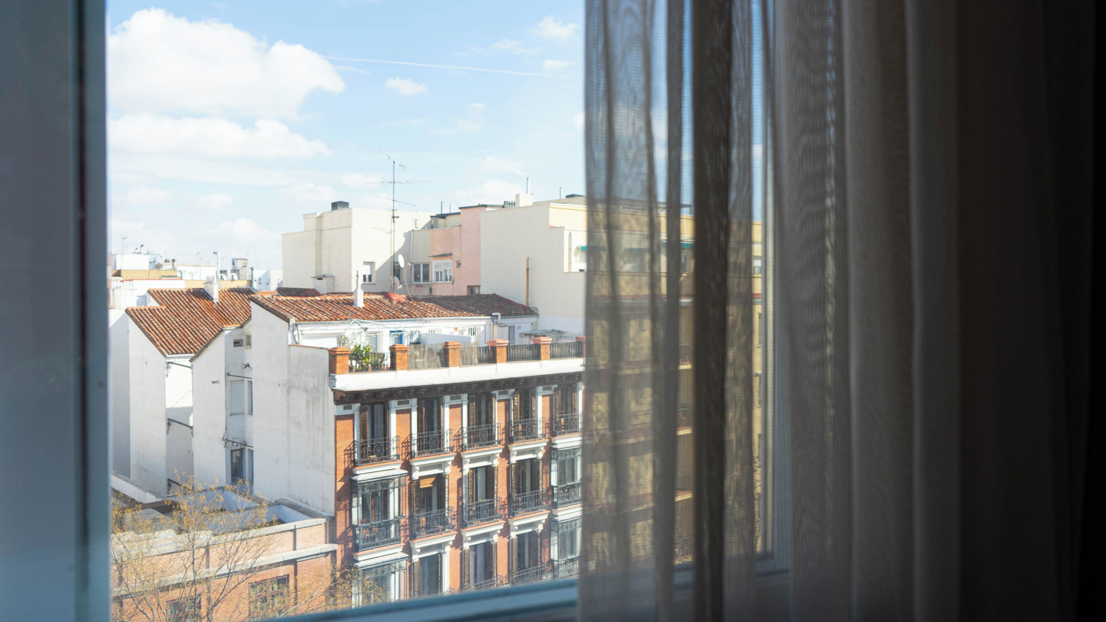A view through a window with curtains, featuring a building with a tiled roof and multiple balconies.