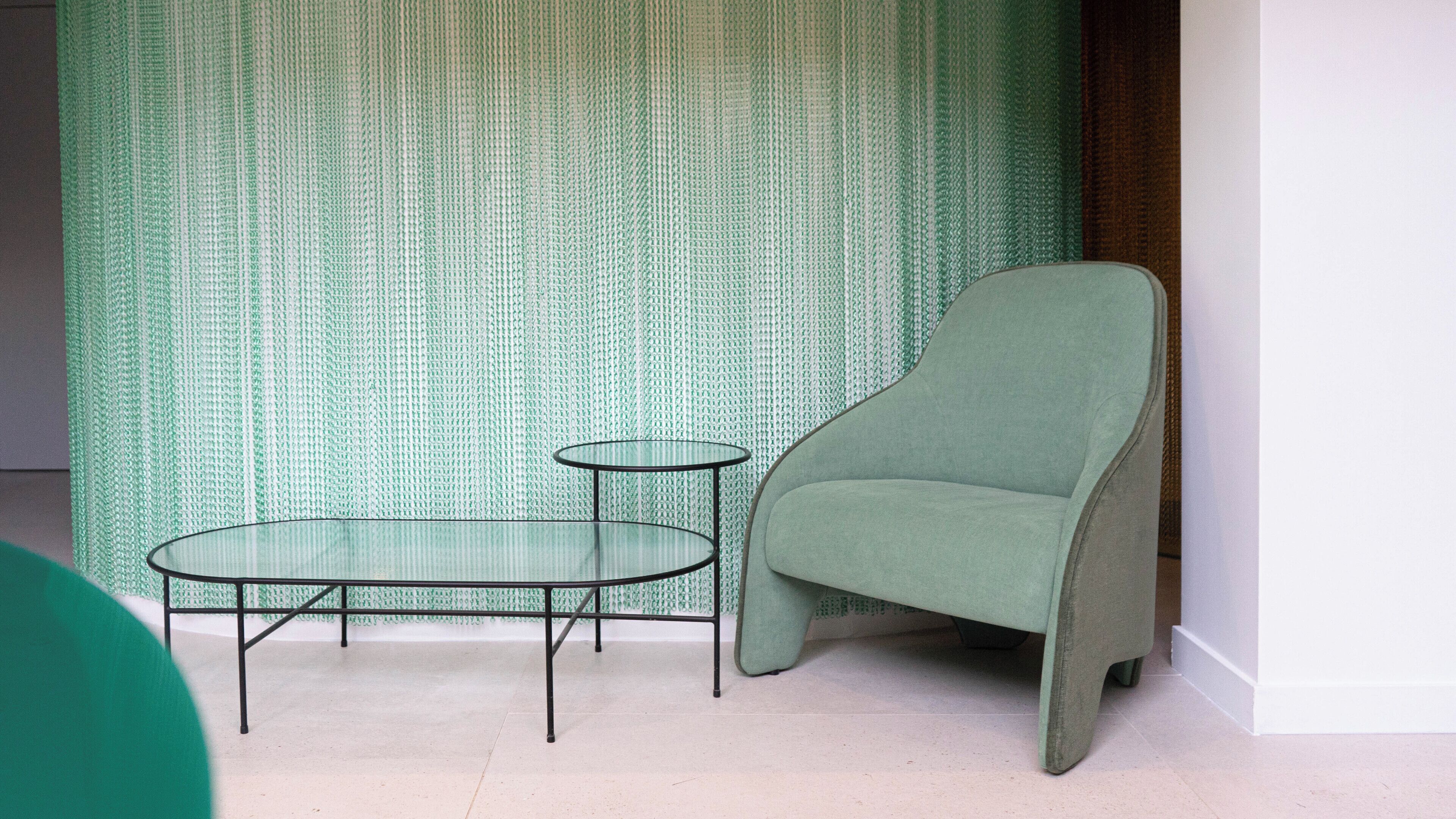 Una habitación con pared de diseño verde, un sillón verde y una mesita de cristal con patas metálicas.