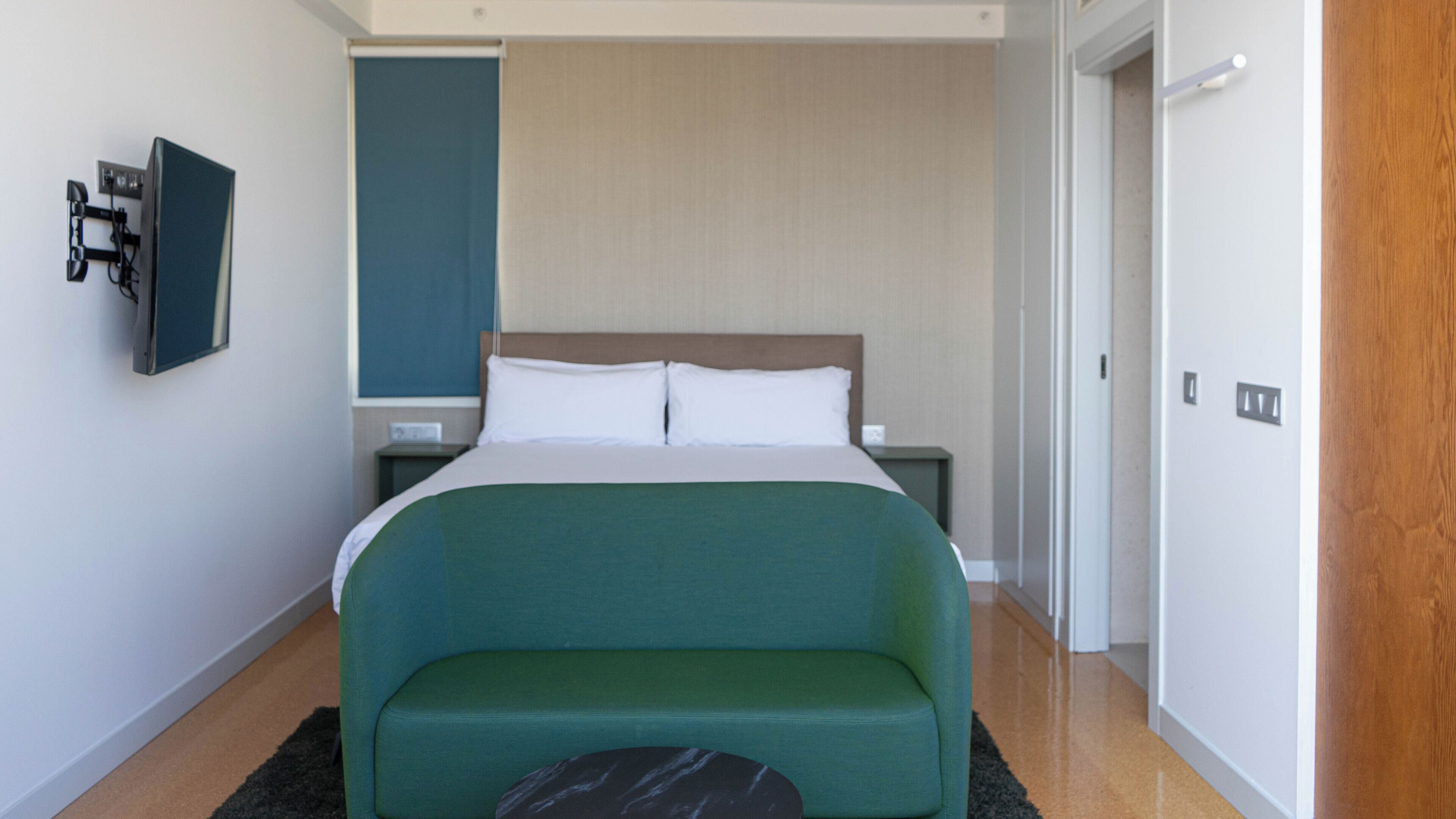 A modern hotel room with a large bed, a green sofa, a flat-screen TV mounted on the wall, and a wooden door.