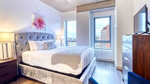 A modern bedroom with a large bed, a city view, and a television. - Luxury Apt Kendall Sq with Amenities (Cambridge MA)