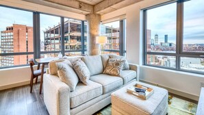 A modern living room with a grey sofa, a small table, and a coffee table. Large windows offer a view of the city skyline and construction sites. - Luxury Apt Kendall Sq with Amenities (Cambridge MA)