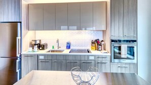 A modern kitchen with grey cabinets, stainless steel appliances, and a central island with a wire basket. - Luxury Apt Kendall Sq with Amenities (Cambridge MA)