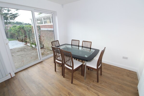 A dining area with a glass table and wooden chairs, a sliding glass door leading to a garden, and a white interior. - Closer to International Ice Bowl & Ulster Hospital (Belfast)