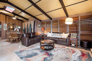 Uma sala de estar ampla, com teto alto, paredes de madeira, sofá de couro, mesa de centro redonda e um espaço de jantar com mobiliário de vime.