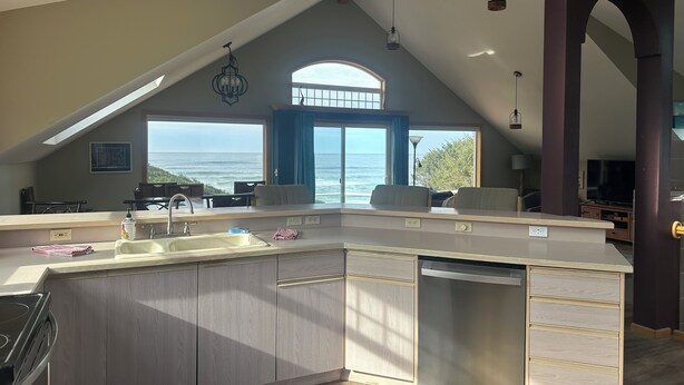 A modern kitchen with a large island, a built-in dishwasher, and a view of the ocean through a large window. - Soaring Seabird - Yachats Ocean View with Hot Tub (Yachats)