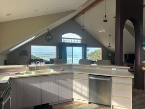 A modern kitchen with a large island, a built-in dishwasher, and a view of the ocean through a large window. - Soaring Seabird - Yachats Ocean View with Hot Tub (Yachats)