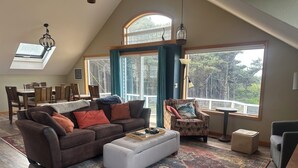 A spacious living room with a high ceiling, a dining area with wooden furniture, and large windows offering a view of the outdoors. - Soaring Seabird - Yachats Ocean View with Hot Tub (Yachats)