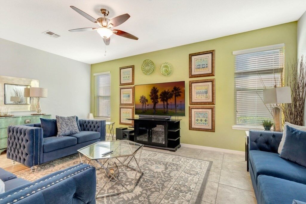 A living room with a blue sofa set, a glass coffee table, a ceiling fan, and framed artwork on the wall.