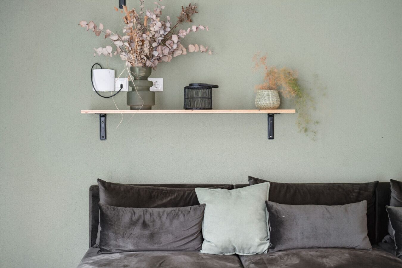 Una habitación con un sofá gris, dos almohadas, un florero con flores secas y un estante con una lámpara y una cesta.