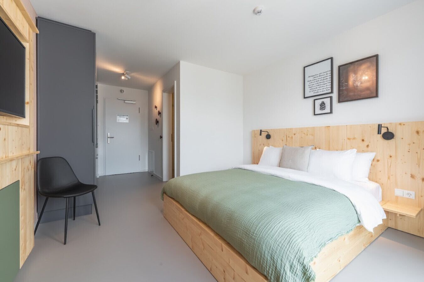 Un dormitorio con cabecera de madera, colcha verde, silla negra y dos cuadros en la pared.