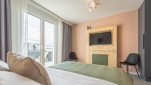 Habitación de hotel moderna con una cama grande, un televisor montado en un panel de madera, dos sillones y vistas al balcón.