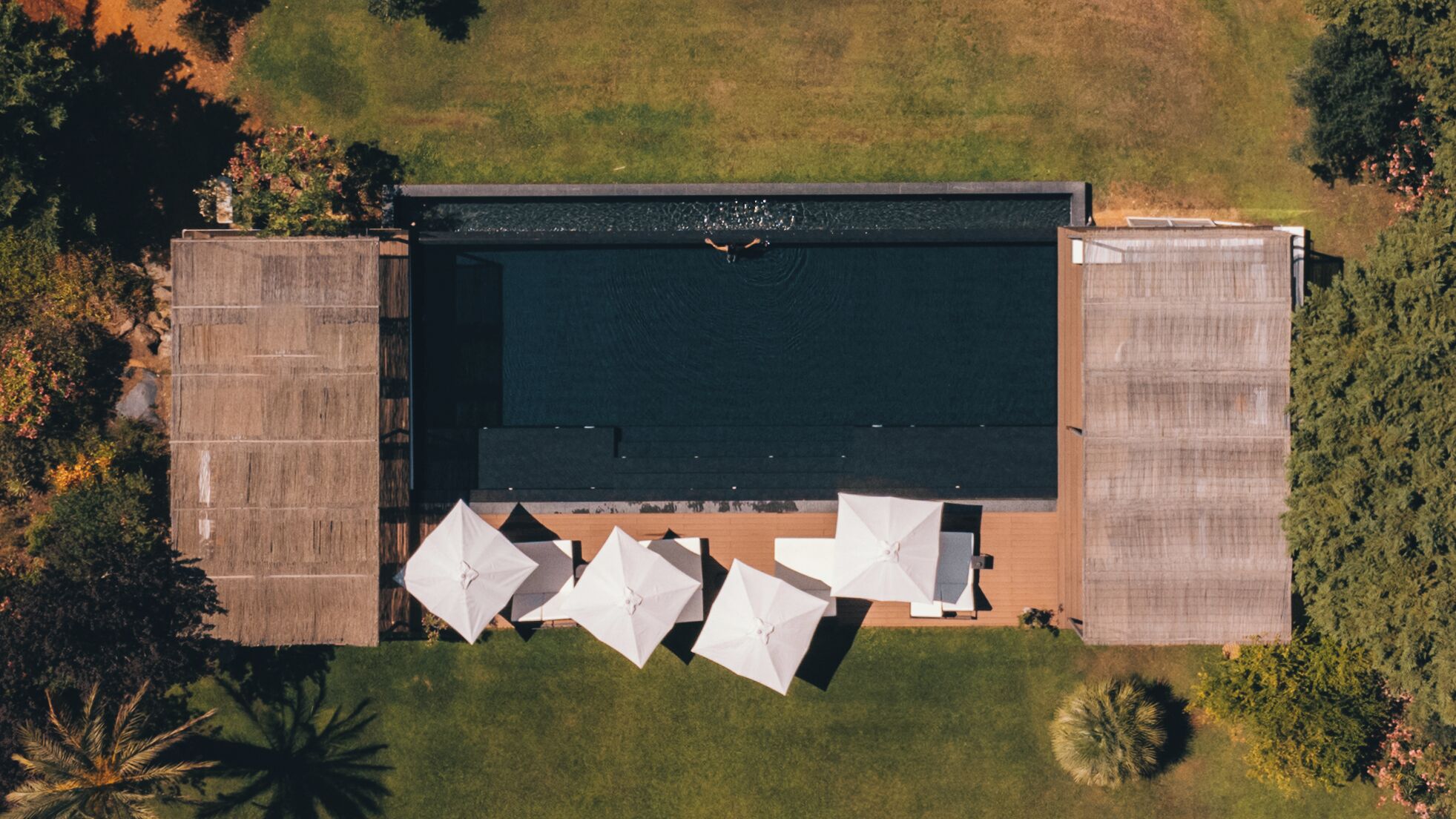 Une vue aérienne d’une piscine rectangulaire où une personne nage, entourée d’une terrasse agrémentée de parasols.