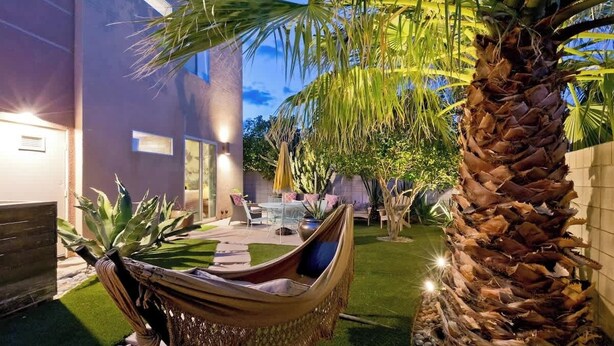 A hammock in a well-lit outdoor area with a palm tree, a modern building, and a patio with outdoor furniture.