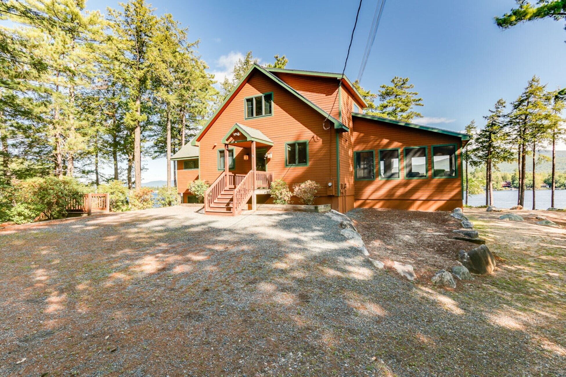A two-story wooden cabin with a front porch, a gravel driveway, and a forested backdrop.