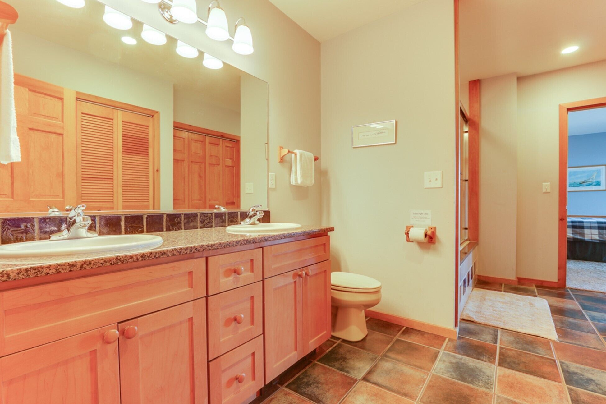 A bathroom with two sinks, a toilet, and a mirror. There are wooden cabinets and a tiled floor.
