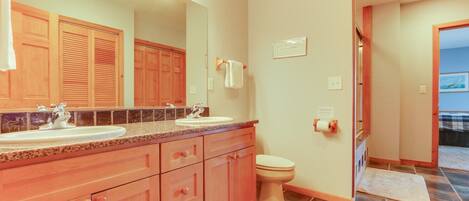 A bathroom with two sinks, a toilet, and a mirror. There are wooden cabinets and a tiled floor.