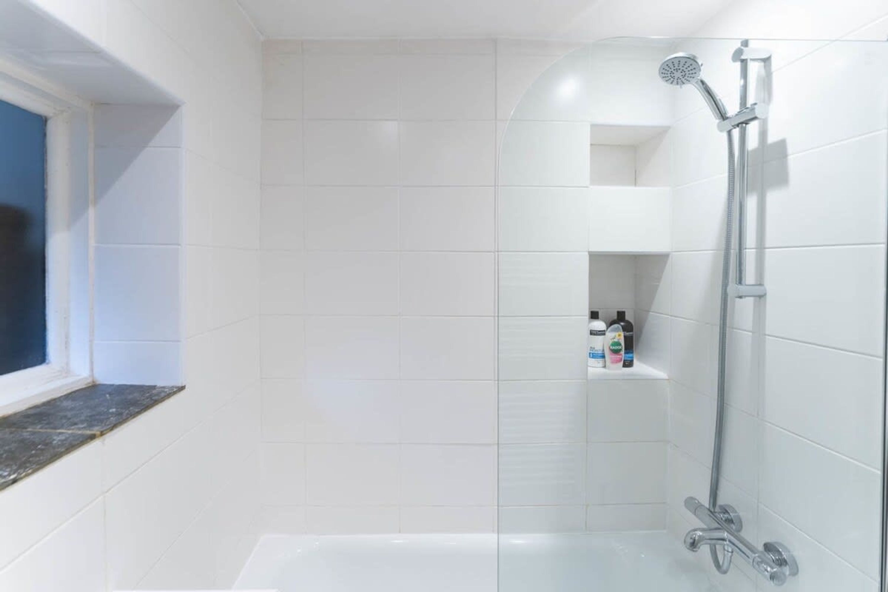 Une salle de bain avec des carreaux blancs, une baignoire, une douche avec un pommeau de douche amovible et une fenêtre à cadre foncé.