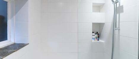 Une salle de bain avec des carreaux blancs, une baignoire, une douche avec un pommeau de douche amovible et une fenêtre à cadre foncé.