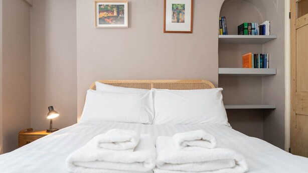 A neatly made bed with white linens and folded towels, a bedside lamp, and a bookshelf with books. - Host Stay Waterloo Apartment (Hove)