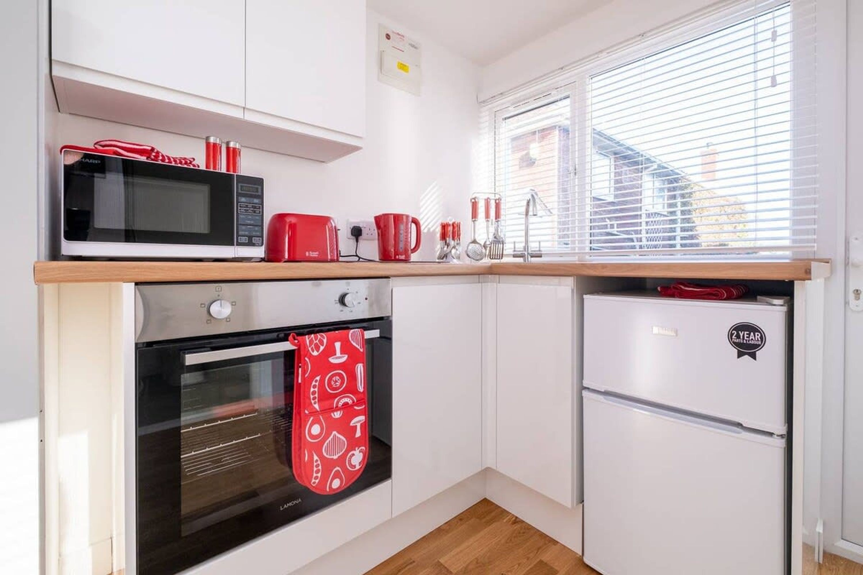 A compact kitchen with a microwave, toaster, and refrigerator, a window with blinds, and a hanging red oven mitt.