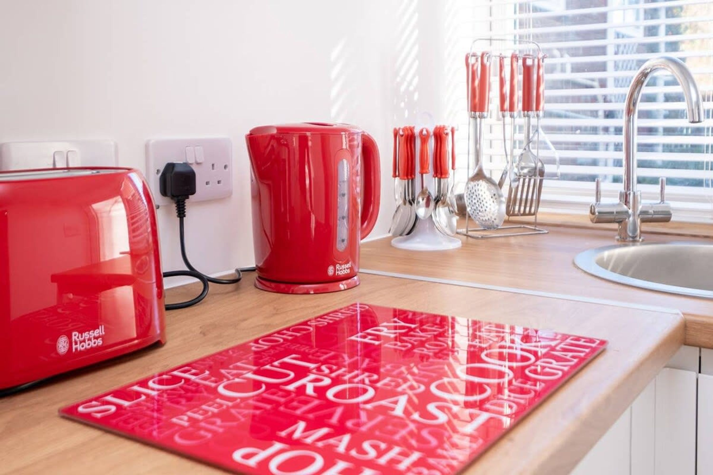 A kitchen with a red Russell Hobbs toaster, a red coffee maker, and a red mug on the counter. There are kitchen utensils in the background and a red placemat with food-related words.