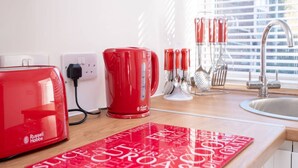 Une cuisine avec un grille-pain Russell Hobbs rouge, une machine à café rouge et une tasse rouge posée sur le comptoir. On aperçoit des ustensiles de cuisine à l’arrière-plan, ainsi qu’une nappe rouge ornée de mots liés à la nourriture.