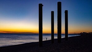 Uma praia com quatro postes verticais e uma pessoa caminhando na orla.