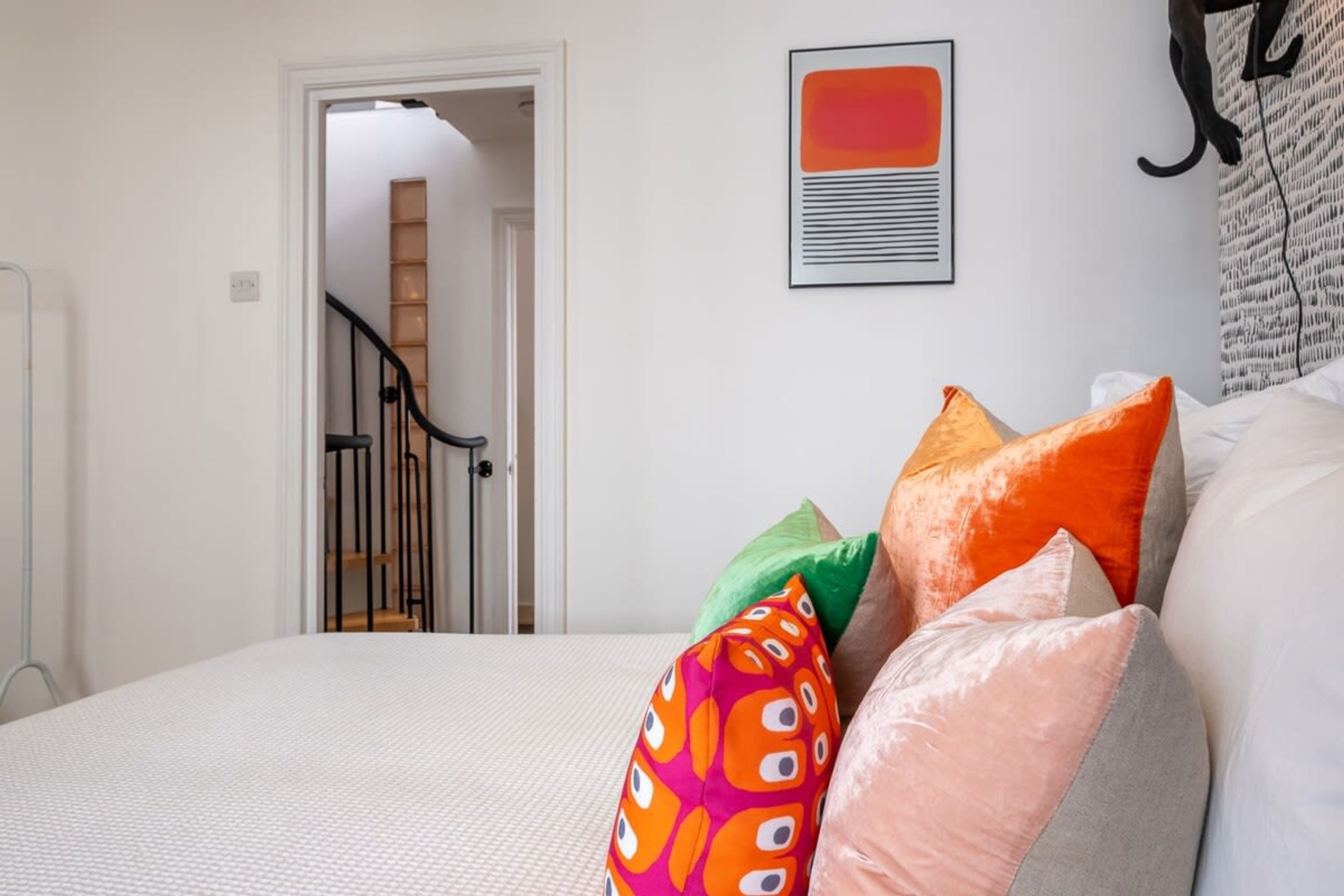 A bedroom with a bed, colorful pillows, a framed artwork, and a staircase leading to another room.