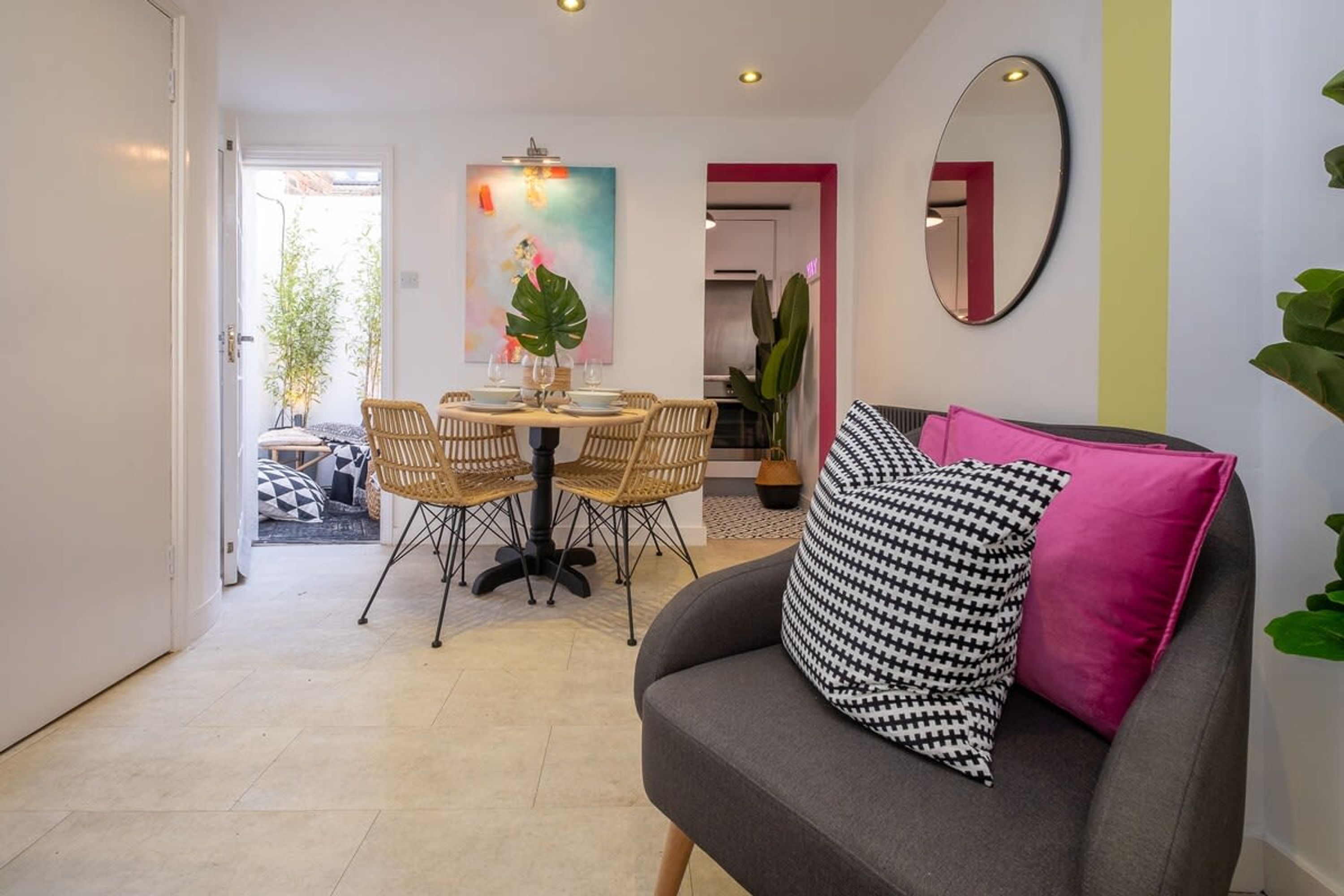 A modern living room with a grey sofa, a round dining table with wicker chairs, and a large mirror on the wall.