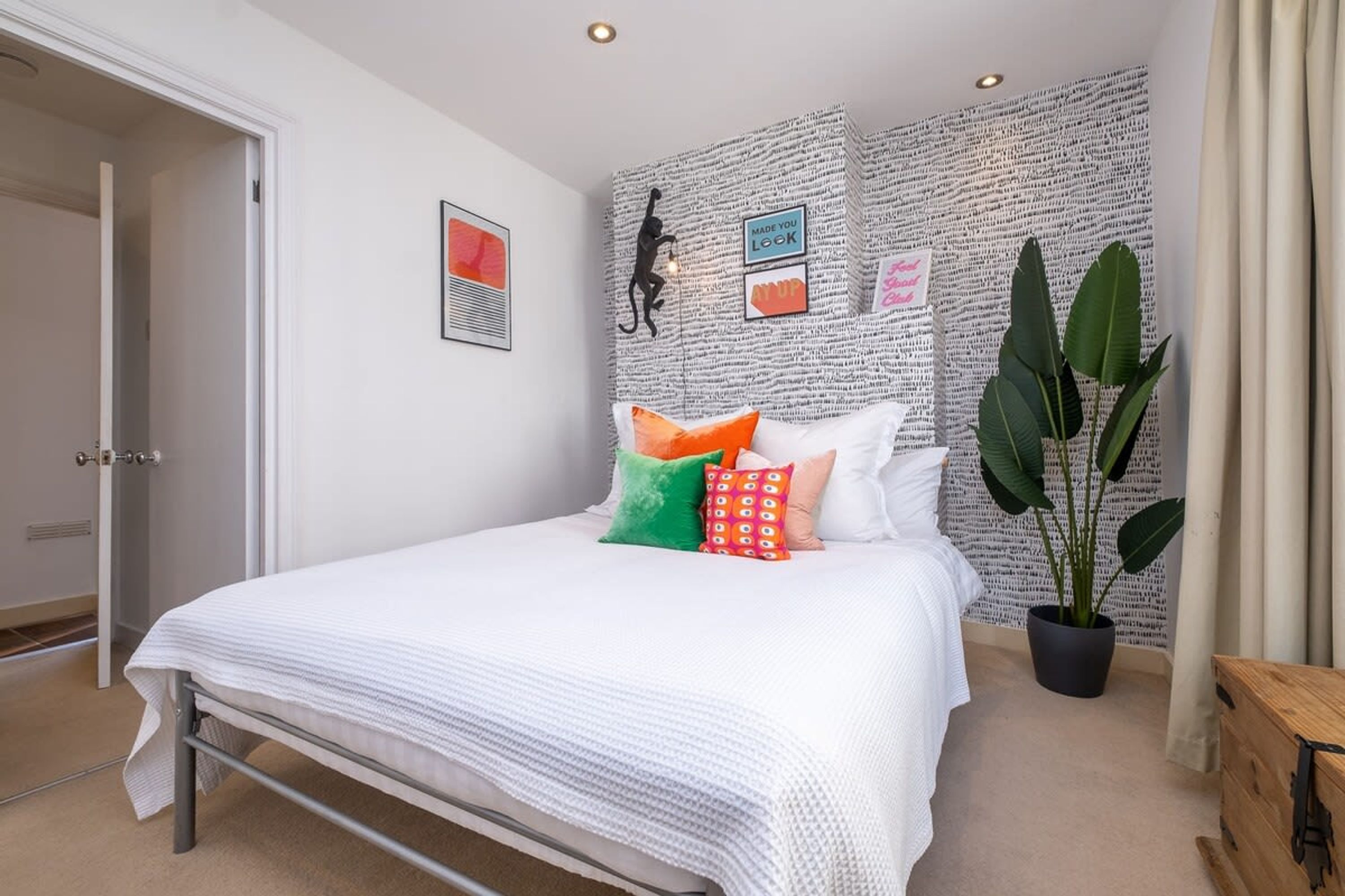 A bedroom with a white bed, colorful pillows, a large plant, and a wall with printed text and a silhouette decoration.