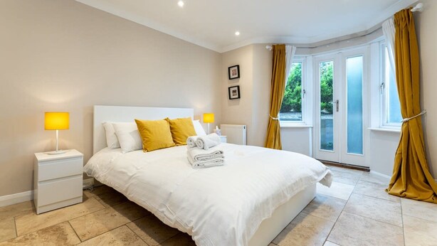A bedroom with a large bed, two bedside tables, a window with curtains, and a view of greenery outside. - Host Stay Seven Dials Apartment (Brighton)