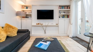 A modern living room with a flat-screen TV mounted on the wall, a sofa with decorative pillows, a coffee table with a book, and a bookshelf filled with books. - Host Stay Seven Dials Apartment (Brighton)