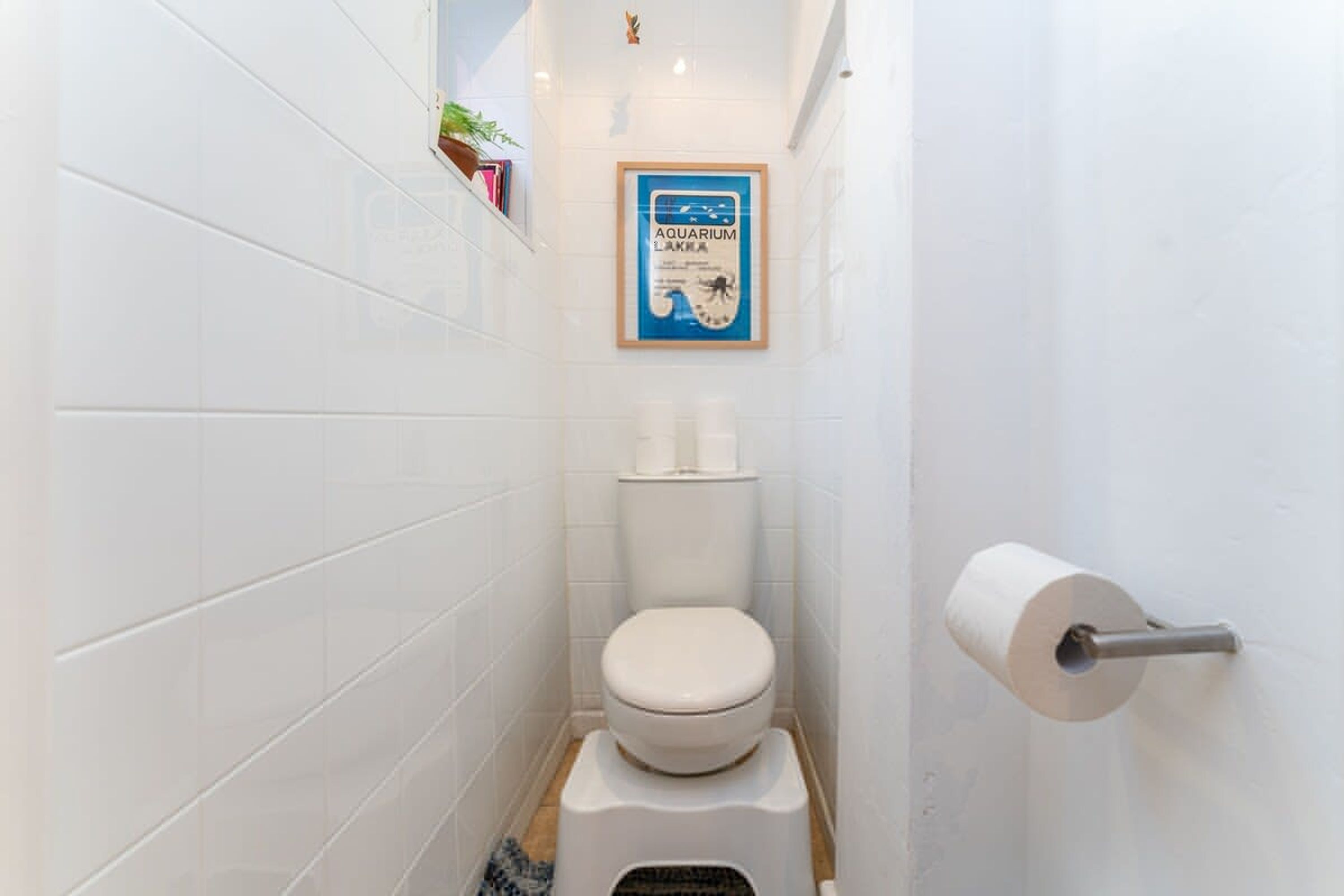 Une petite salle de bain aux murs recouverts de carreaux blancs, avec des toilettes, un rouleau de papier toilette et une affiche encadrée accrochée au mur.