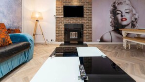 A living room with a brick fireplace, a flat-screen TV, a sofa with orange cushions, a coffee table, and a lamp.
