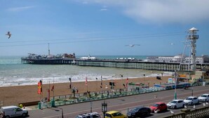 A coastal scene with a pier extending into the sea, a beach with flags, and a road with parked cars.
