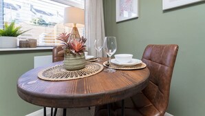 A dining area with a round wooden table, a brown leather chair, a woven placemat, a vase with plants, a wine glass, and a white bowl. - Host Stay Round Hill Apartment (Brighton)