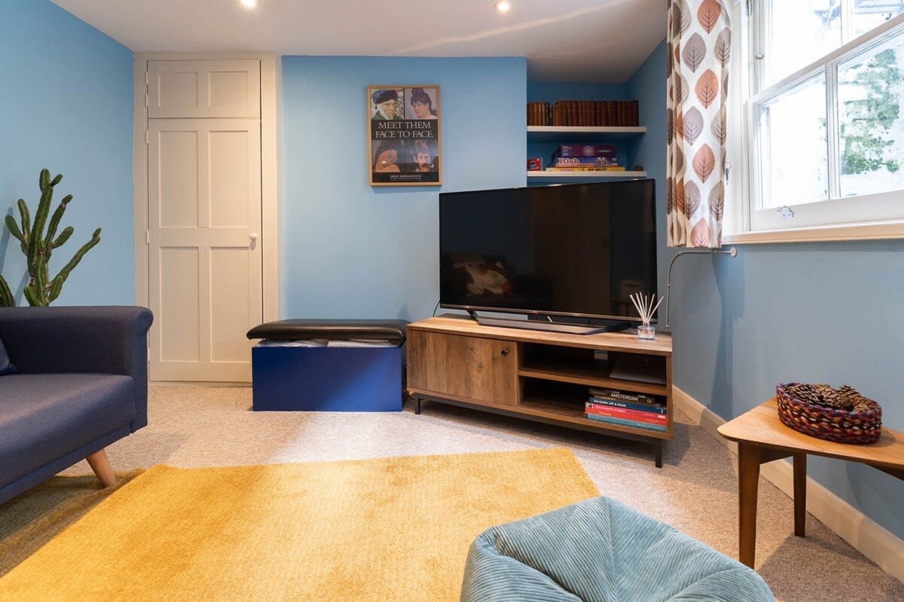 A living room with a blue sofa, a yellow rug, a television on a wooden stand, and a bookshelf.