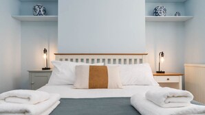 Ein ordentlich bezogenes Bett mit weißen und braunen Kissen, zwei Nachttische mit Lampen und ein Stapel weißer Handtücher auf dem Boden.
