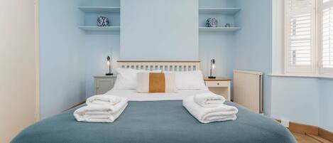 A bedroom with a bed, two bedside tables, a nightstand, a radiator, and a window with blinds.