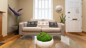 Ein Wohnzimmer mit einem beigen Sofa, einem Glastisch mit einem Grünmoos-Dekor, einer hohen, violetten Blumenkomposition und einer weißen Tür mit einem Schild.