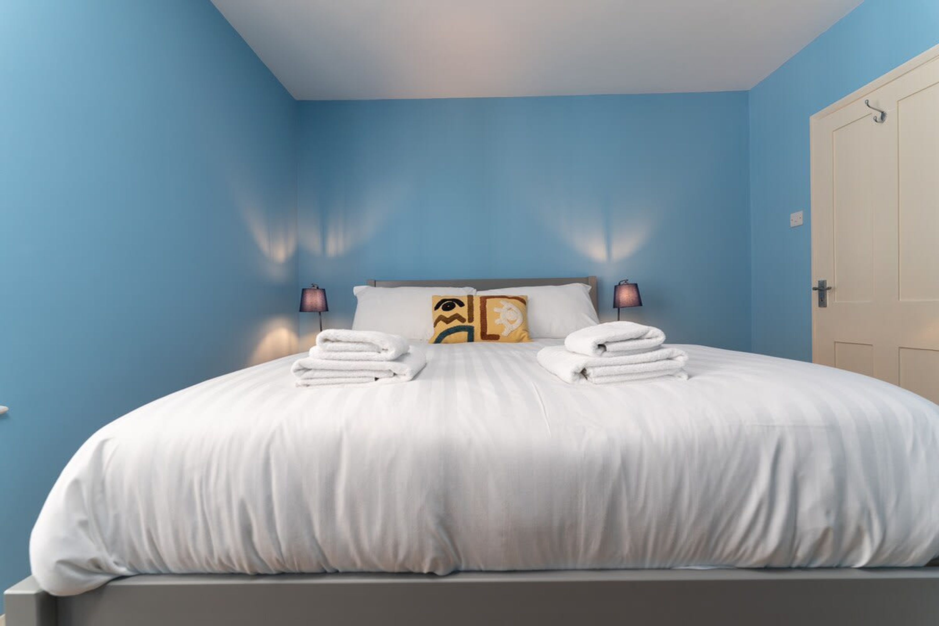 A neatly made bed with white linens and two pillows, a bedside lamp, and a door to the right.